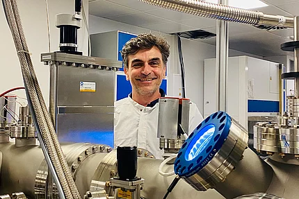 Man in a lab coat smiling behind stainless steel scientific equipment, including a circular blue-lit component in the foreground, in a research lab setting.