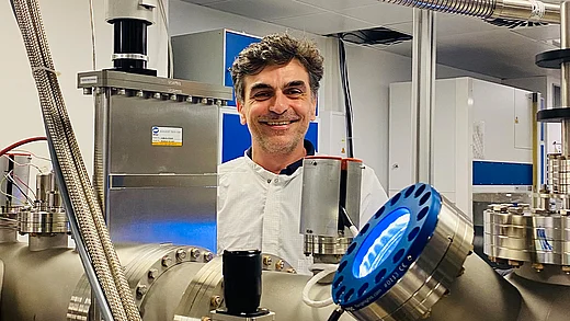Man in a lab coat smiling behind stainless steel scientific equipment, including a circular blue-lit component in the foreground, in a research lab setting.