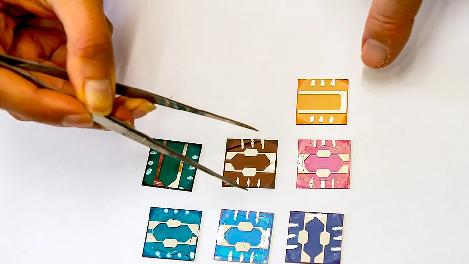 Close-up of a researcher's hands using tweezers to handle small square organic solar cell samples. Six devices in various colours including teal, brown, pink, and blue are arranged on a white surface, each displaying a distinctive patterned metallic electrode structure.