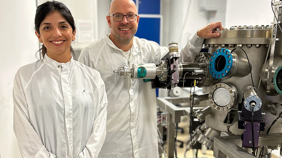 Two researchers in white cleanroom suits smile at the camera in a lab; the man on the right rests his hand on a large stainless-steel ultra-high-vacuum chamber fitted with viewports and flanges.