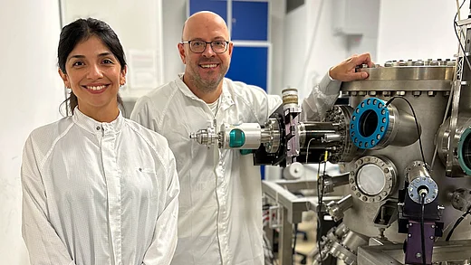 Two researchers in white cleanroom suits smile at the camera in a lab; the man on the right rests his hand on a large stainless-steel ultra-high-vacuum chamber fitted with viewports and flanges.